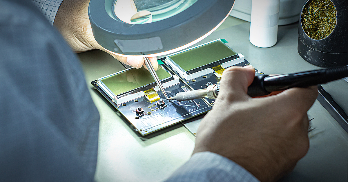 An AKSO technician assembling an instrument circuit board under a magnifier lamp